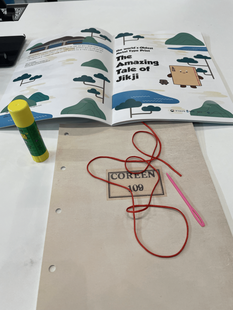 A table setup featuring an open booklet titled 'The Amazing Tale of Jikji', a glue stick, a needle, and red thread, alongside a cover labeled 'COREEN 100'. Ready to sew the binding!