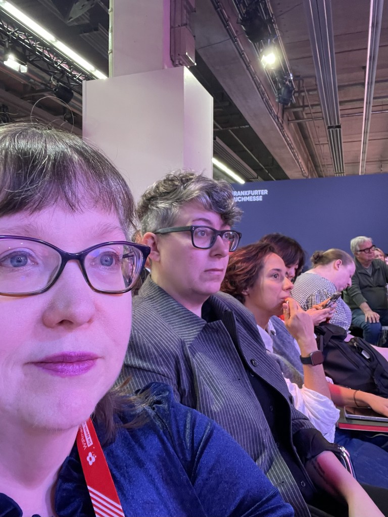 A group of people seated in an audience at the Frankfurt Book Fair, with one person in the foreground taking a selfie.